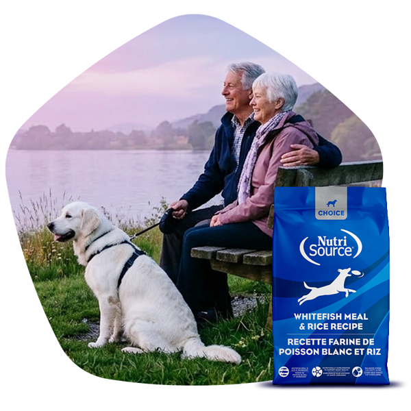 Older couple with a dog sitting on a bench by a scenic lake, NutriSource® Choice dog food bag in the foreground