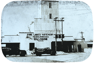 Vintage black and white photo of Pine Lakes Hatchery Feed & Produce building with trucks and equipment outside