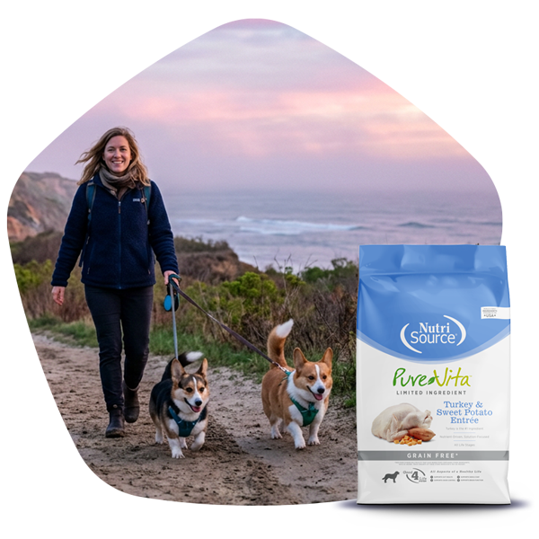 Woman walking two dogs on a trail with a scenic background, NutriSource® PureVita dog food bag in the foreground